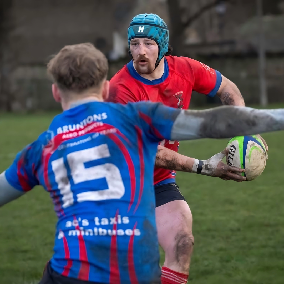 Two rugby players in action on a grass field, one in a red jersey and blue helmet, the other in a blue jersey with visible text.